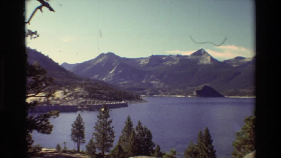 1981 8mm frame showing a lake below tree-covered mountains, with conifers in the foreground and a dam or bridge-like structure near shore.