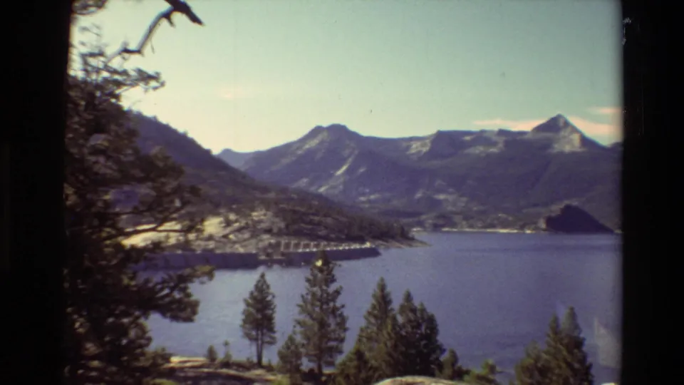 1981 8mm frame of a mountain lake in a wooded overlook, with branches at the edge, a shoreline dam, and layered ridges in the distance.