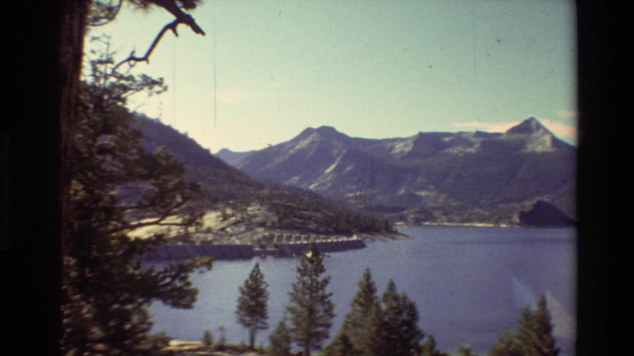 1981 8mm home-movie view of a mountain lake in the British Columbia metadata location, with evergreens and a distant dam or bridge.