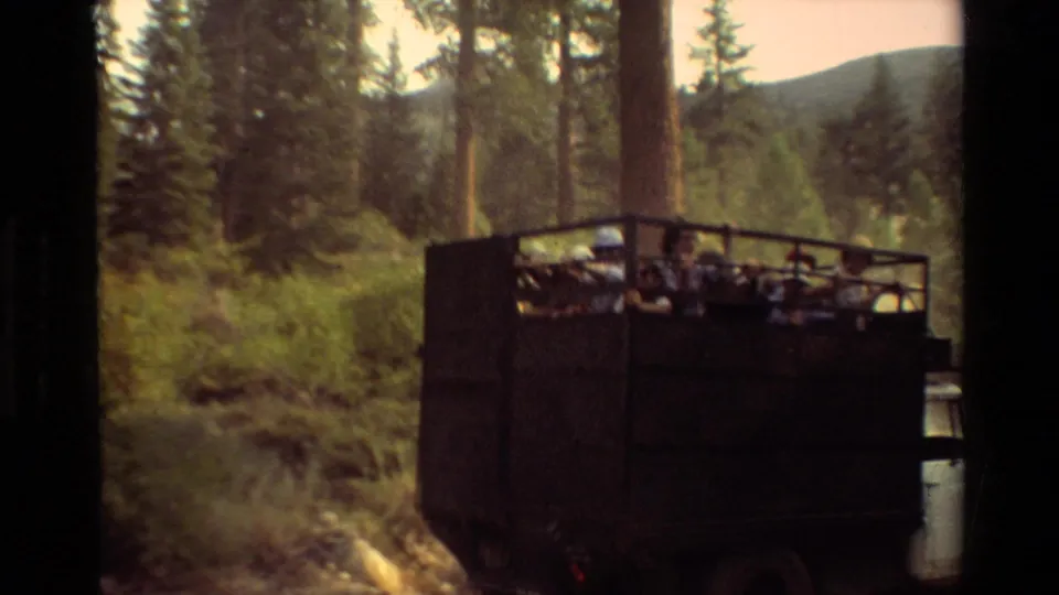 An open-sided truck with seated passengers moves through a wooded mountain road in this 1981 8mm travel scene.