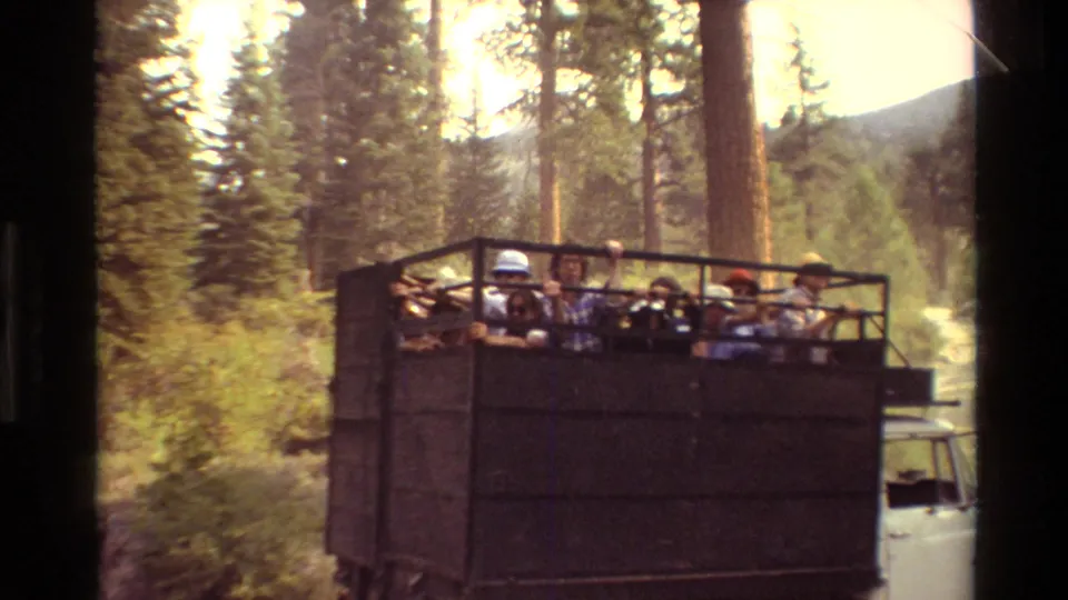Passengers in casual clothing sit in an open truck bed as it passes through dense trees and mountain greenery.