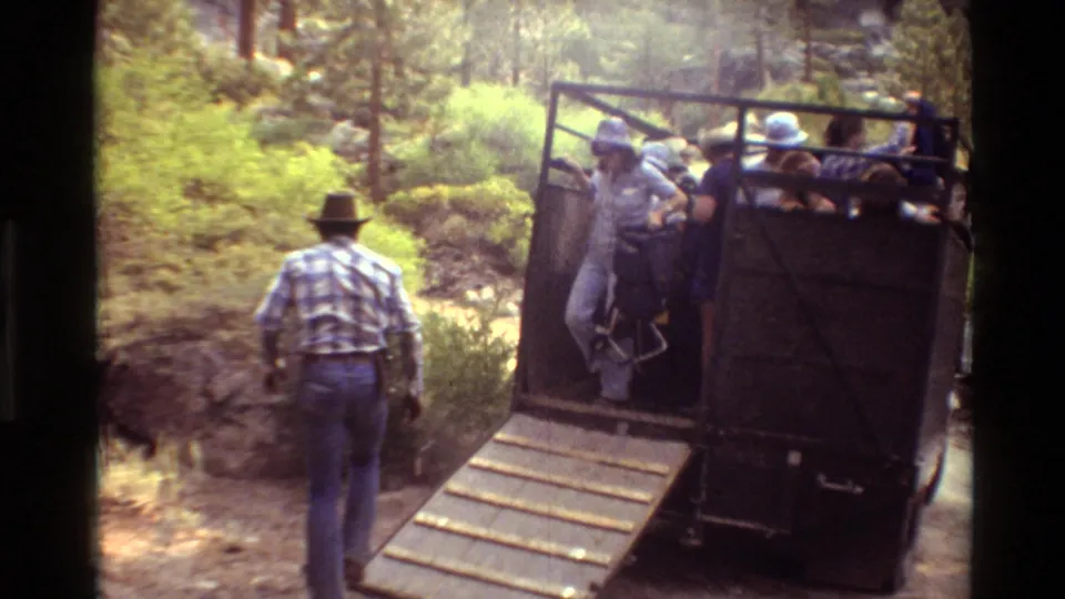 Tourists in hats step down a wooden ramp from the open rear of a truck in a forest setting.