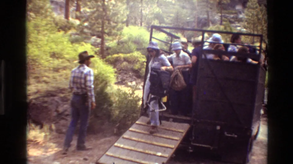 People in casual clothing and hats exit the open tour truck by ramp as a man stands nearby in the forest.