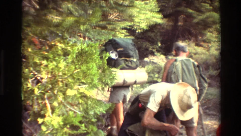 Home-movie view of backpackers walking a dirt trail through pine and brush, carrying bulky camping gear.