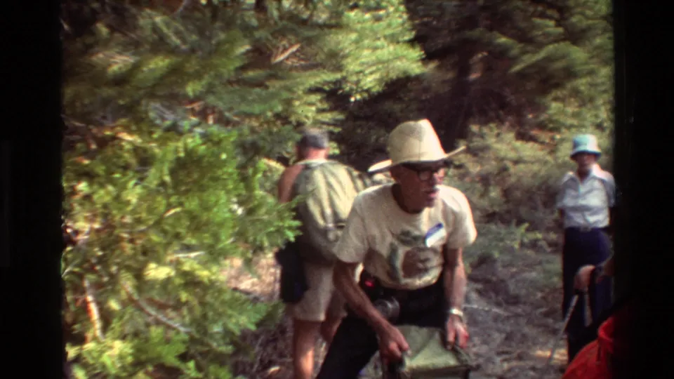 1981-style 8mm frame of backpackers on a wooded trail, including a foreground man in a brimmed hat carrying gear.