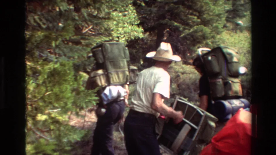 Hikers in casual outdoor clothing move through a wooded trail area with large backpacks and carried gear.