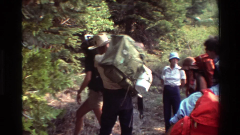 Backpackers with bulky packs pass close to the camera while other hikers stand farther along the wooded path.
