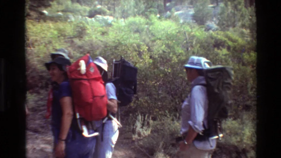 Backpackers in casual outdoor clothing move along a brush-lined trail in metadata-listed British Columbia, with several faces partly visible.