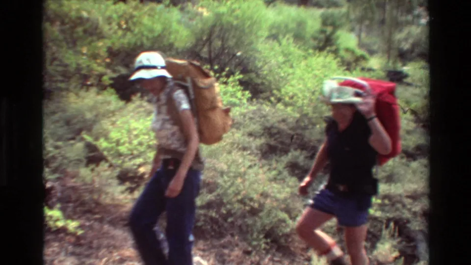 Two backpackers, one with a tall tan pack and one with a red pack, walk through a sunlit wooded trail in the 8mm home-movie sequence.
