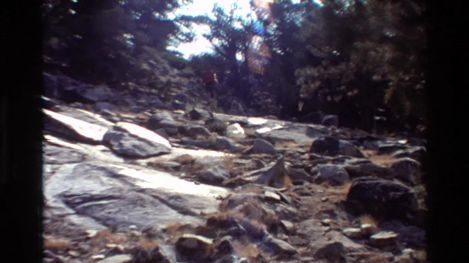 A rocky section of wooded trail appears in bright sunlight, with a distant backpacker partly visible near the upper path.