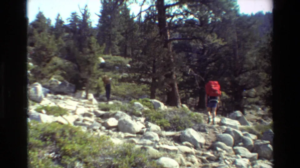 Two backpacked hikers appear on a rocky forest trail in 1981 8mm footage, one carrying a large red pack.