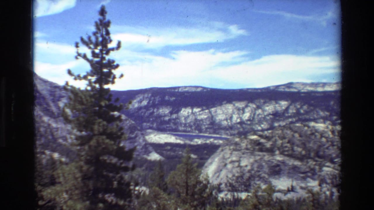 1981 8mm home-movie view of a pine tree before rocky mountain ridges, forested valley, and blue sky with clouds.