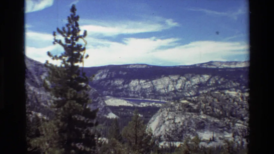 Handheld 8mm landscape view of a tall pine tree against distant rocky mountains, forest, and a clouded blue sky.