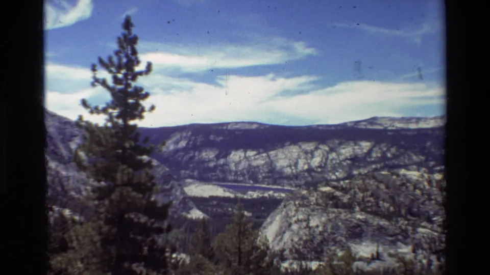 Vintage 8mm scenic overlook showing a pine tree in front of rocky mountain slopes, a valley floor, and bright cloud bands.