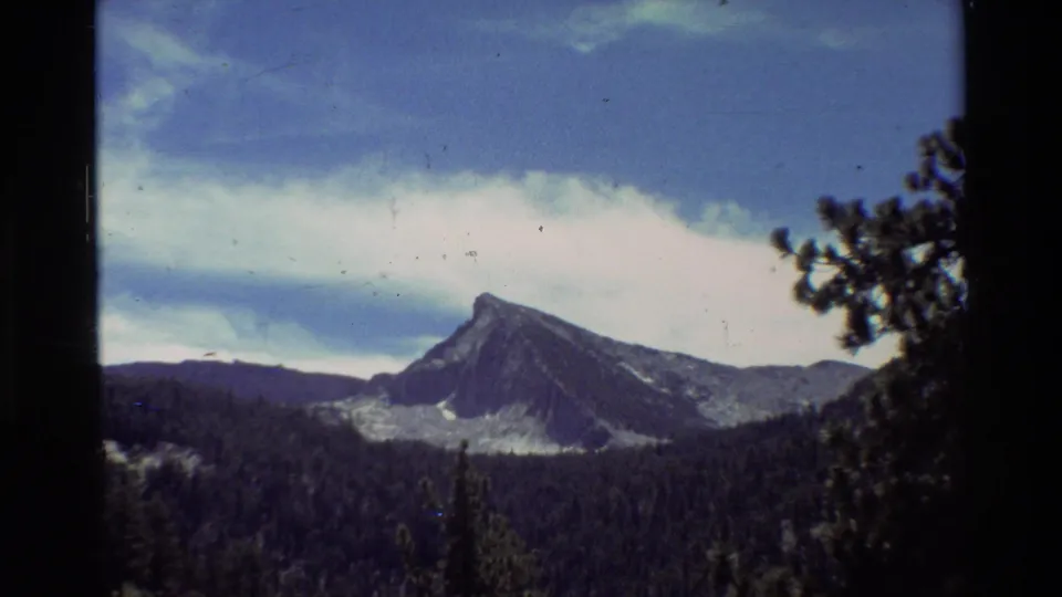 1981 8mm landscape frame with a pointed mountain peak rising beyond dark conifer forest beneath layered clouds.