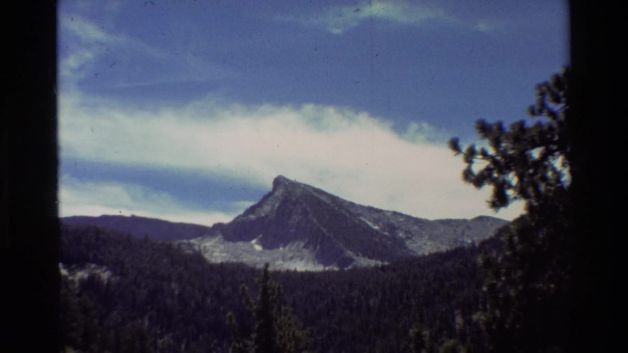 1981 8mm-style mountain view with a rocky peak rising above evergreen forest under a broad blue sky, based on supplied metadata.
