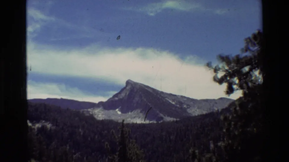 Mountain landscape from supplied 1981 8mm footage, with a distant rocky peak, evergreen forest, and cloud bands across a blue sky.