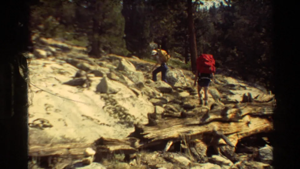 Two distant hikers with large backpacks walk over rocky forest terrain in 1981 home-movie footage from the supplied location.
