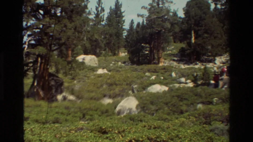 Listed British Columbia trail footage shows a rocky forest slope with pine trees and a distant hiker carrying red gear at right.