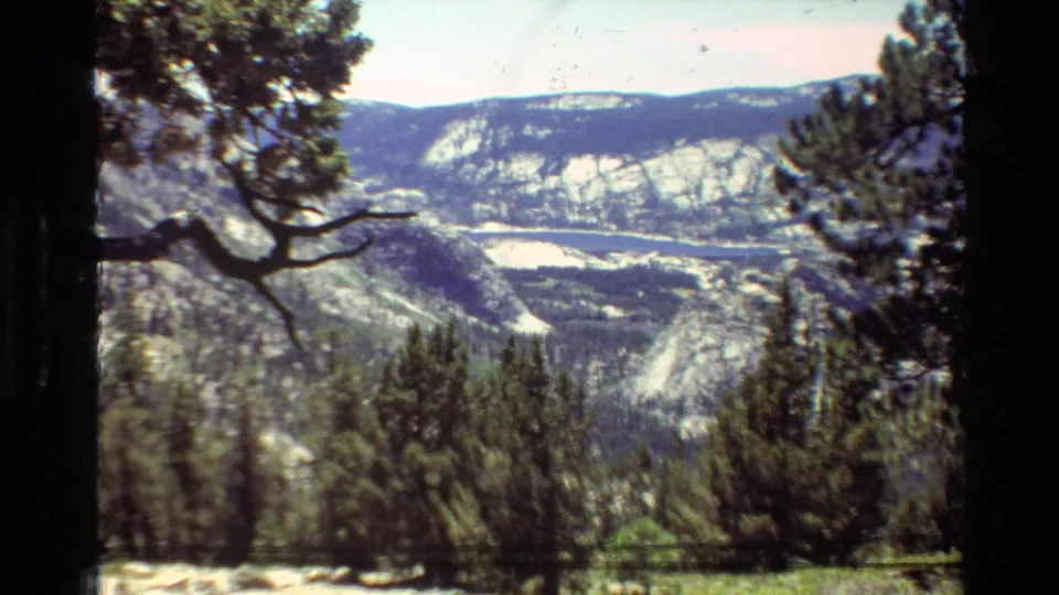 Vintage 8mm frame of a mountain valley seen through pines, with rocky pale slopes and dense forest below.