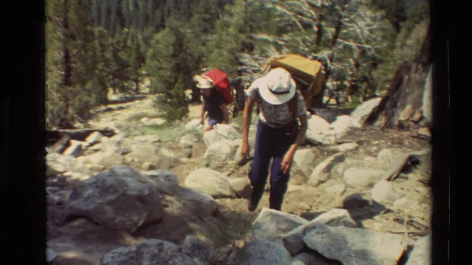 Two hikers on a rocky mountain trail, one in a white hat with a large pack and another with a red pack, in 1981 home-movie footage.
