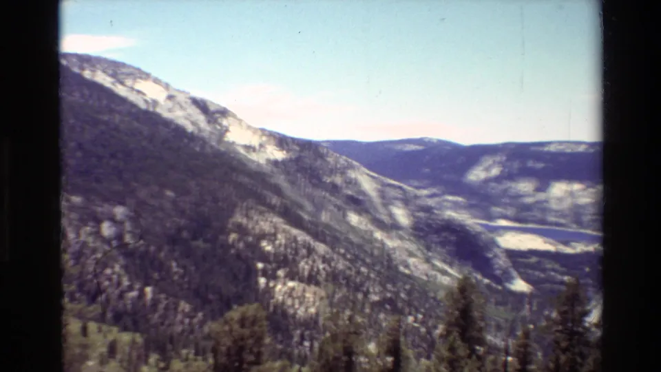 The final 8mm frame opens to a wide mountain panorama with forested slopes, exposed pale rock, distant ridges, and a valley below.