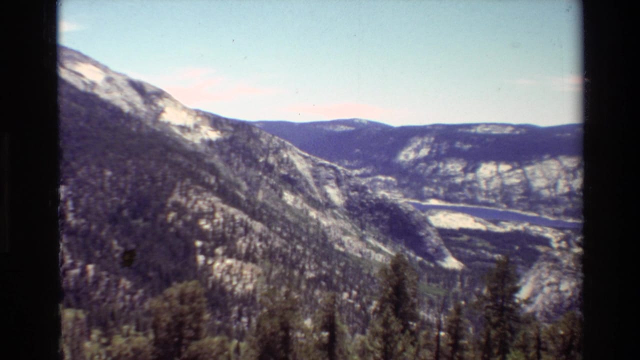 Mountain valley with forested slopes, distant water, pale sky, and dark film border.