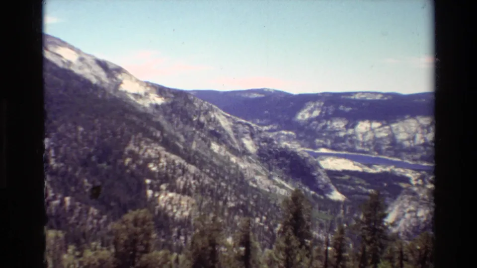 1981 8mm frame of a British Columbia mountain valley with evergreen slopes, distant water, pale sky, and visible film-edge shadow.