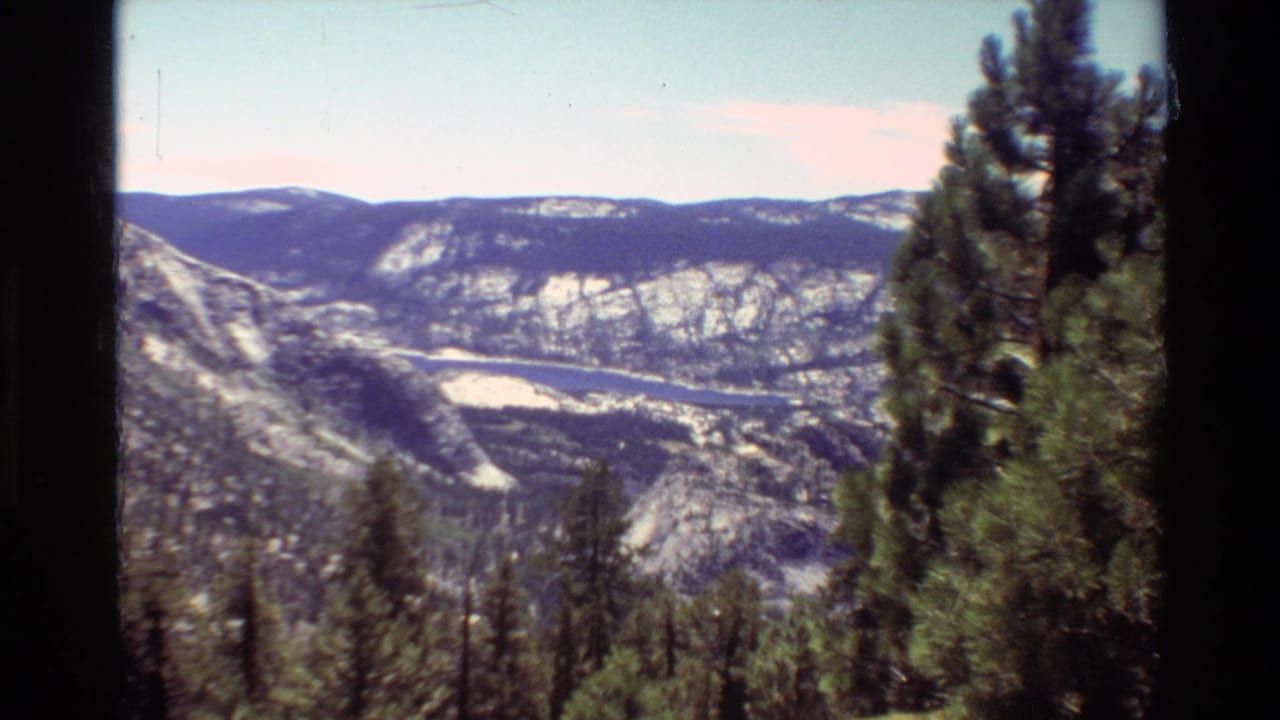 1981 8mm-style mountain view showing evergreen trees, rocky slopes, and a distant valley under a light blue sky.