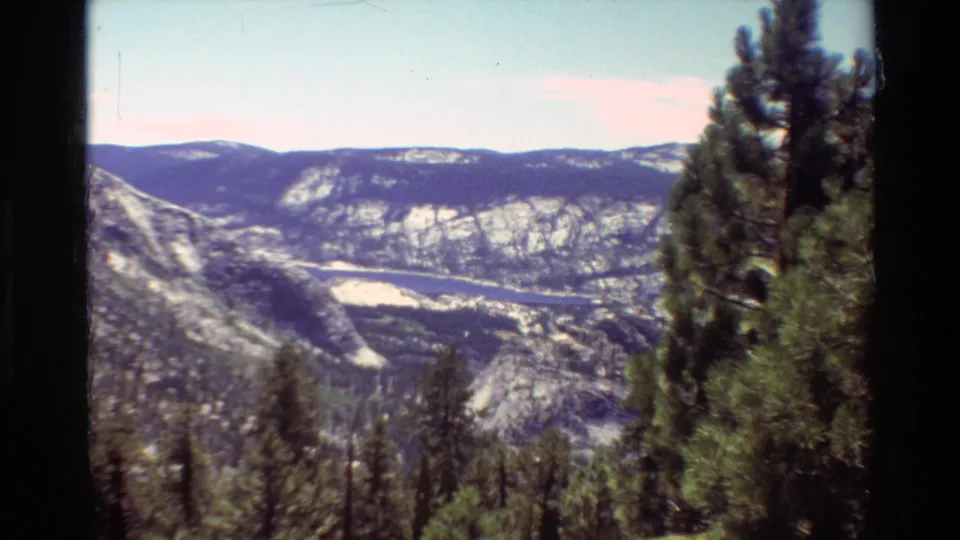1981 8mm-style mountain view showing evergreen trees, rocky slopes, and a distant valley under a light blue sky.
