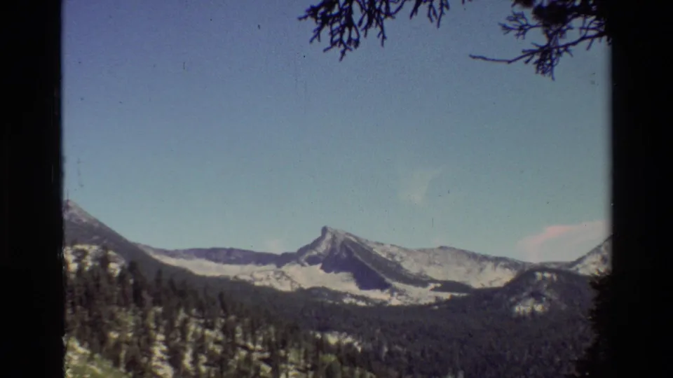 A pointed snow-covered peak stands beyond dark forested hills in vintage 8mm mountain footage from the supplied 1981 reel.