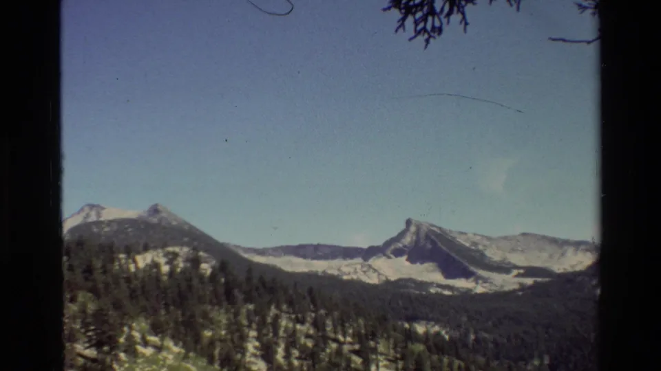 Wide alpine panorama showing snow-streaked ridges and dense evergreen forest in archival 8mm travel footage.