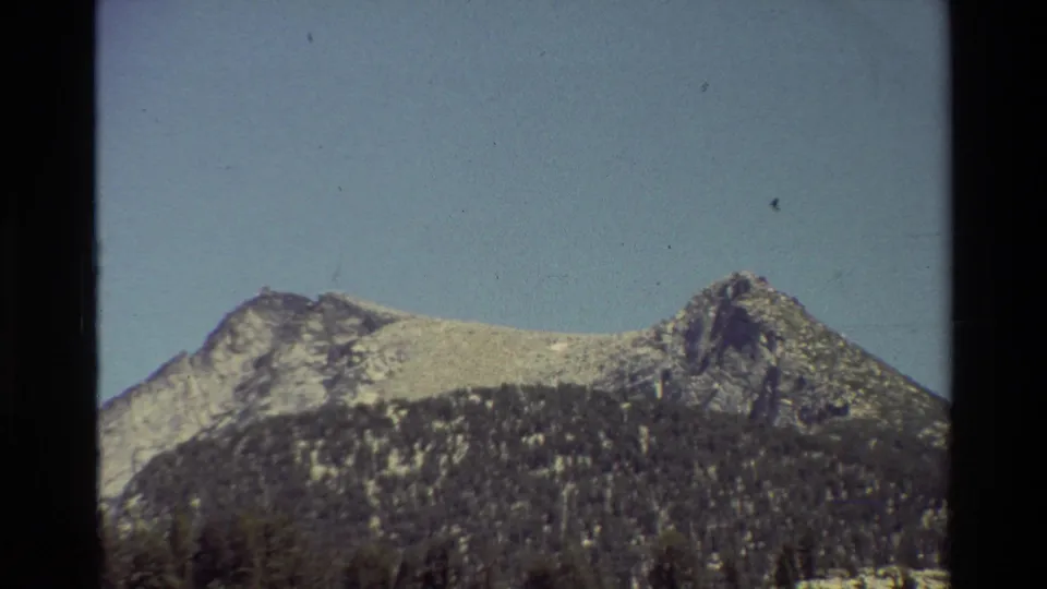 Closer 8mm view of a rugged, snow-dusted mountain face rising above dense conifer forest under a clear blue sky.