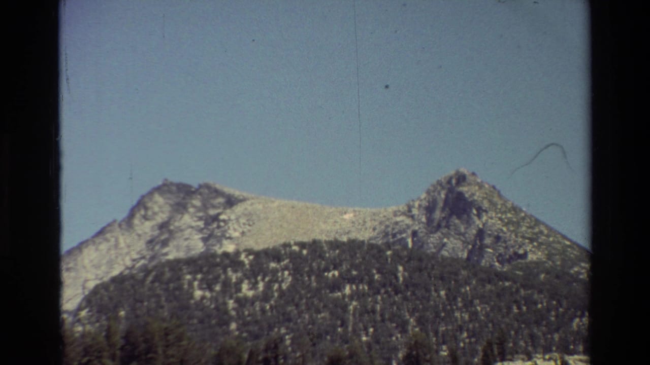 Cataloged 1981 8mm frame of rocky mountain peaks rising above dark forested slopes under blue sky.