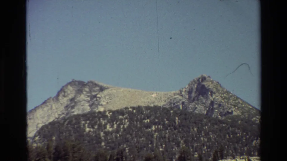 Cataloged 1981 8mm frame of rocky mountain peaks rising above dark forested slopes under blue sky.