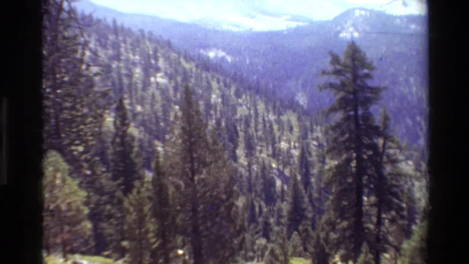 Handheld 8mm view across a conifer-covered mountain valley with distant ridges and bright sky.