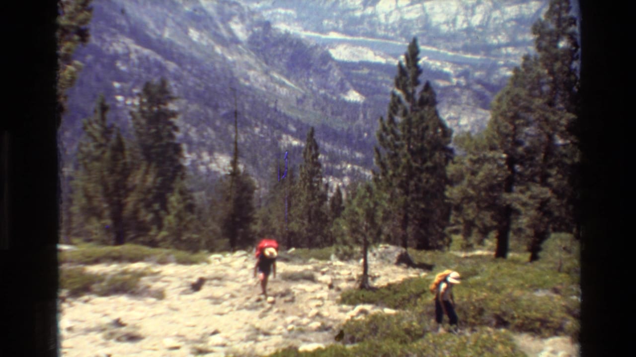 Two adult backpackers, one with a red pack and one with a yellow pack, move uphill on a rocky trail in 1981 mountain footage.
