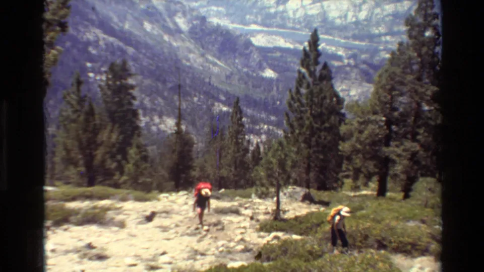 Two adult backpackers, one with a red pack and one with a yellow pack, move uphill on a rocky trail in 1981 mountain footage.