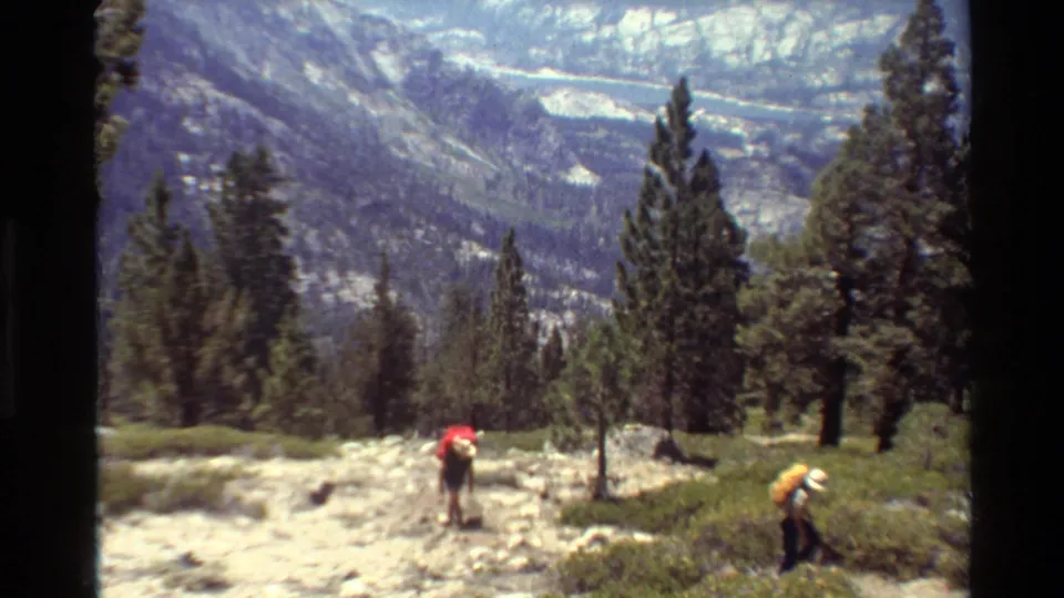 The hikers remain separated on the trail, with red and yellow packs visible against pine trees and a broad mountain backdrop.