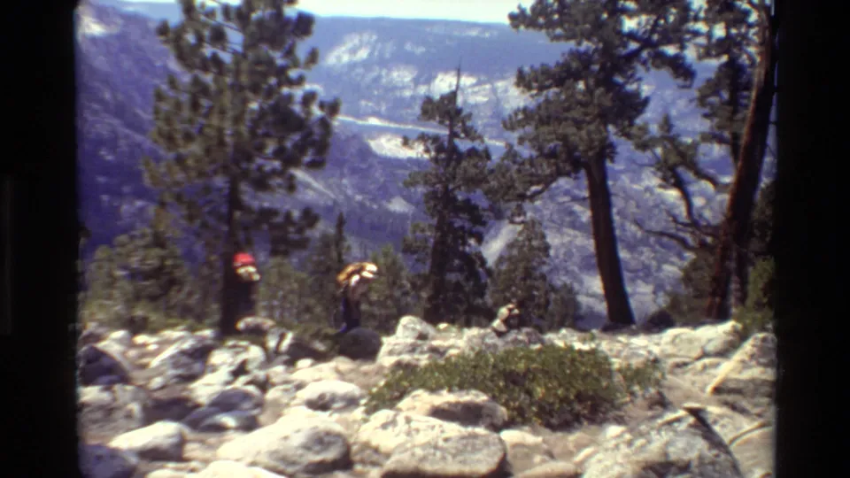 A closing frame shows the two adult backpackers crossing rocky terrain, partly framed by tall trees and distant mountain ridges.