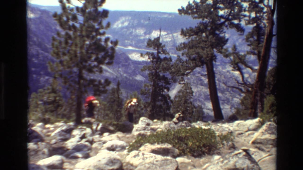 Two backpacked hikers on a rocky mountain trail with pine trees and distant ridges.