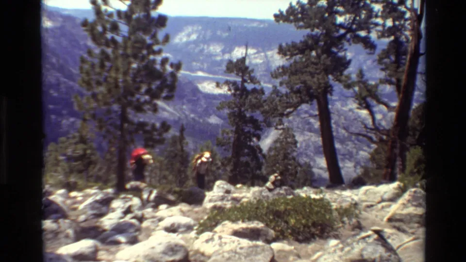 1981 8mm home-movie frame of two hikers with backpacks crossing a rocky mountain trail in the stated British Columbia location.