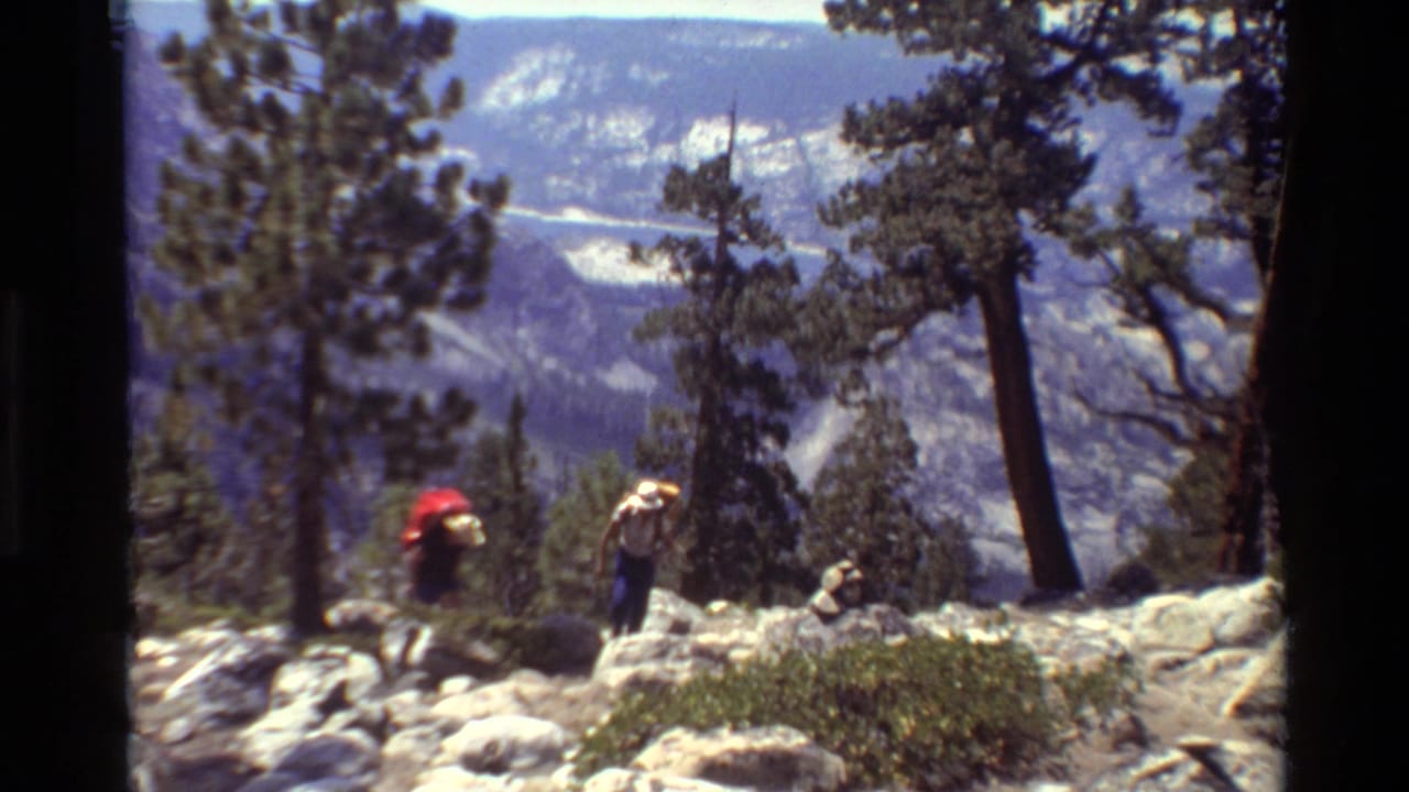 Two hikers, one with a red pack and one in a light hat, walk along a rocky alpine trail in 1981 home-movie footage.
