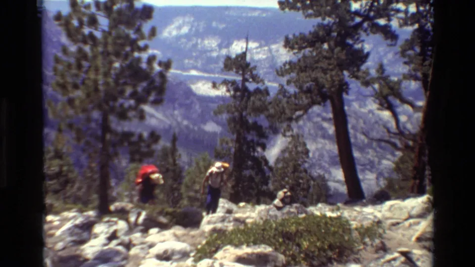 Two hikers, one with a red pack and one in a light hat, walk along a rocky alpine trail in 1981 home-movie footage.