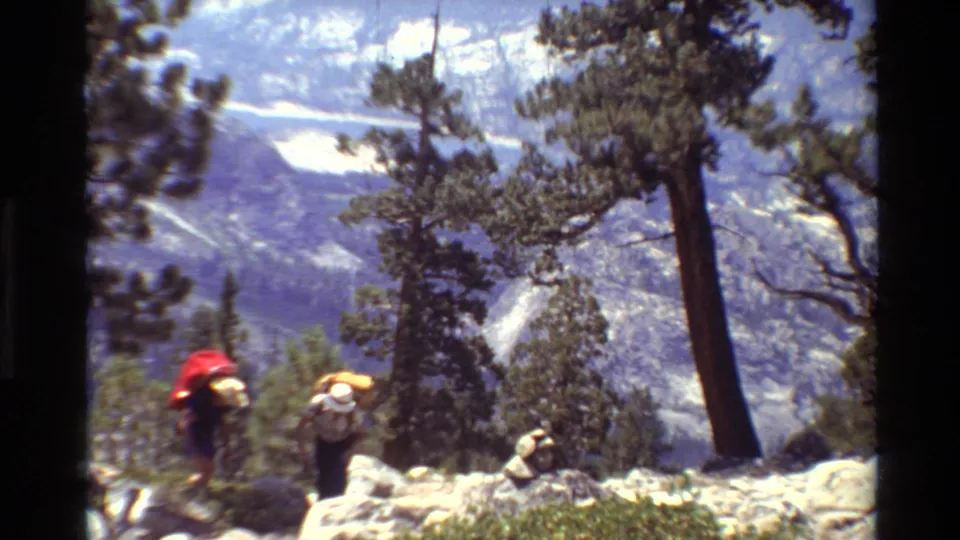 A soft 8mm frame shows two backpacked hikers moving through rocks and brush against a distant mountain landscape.