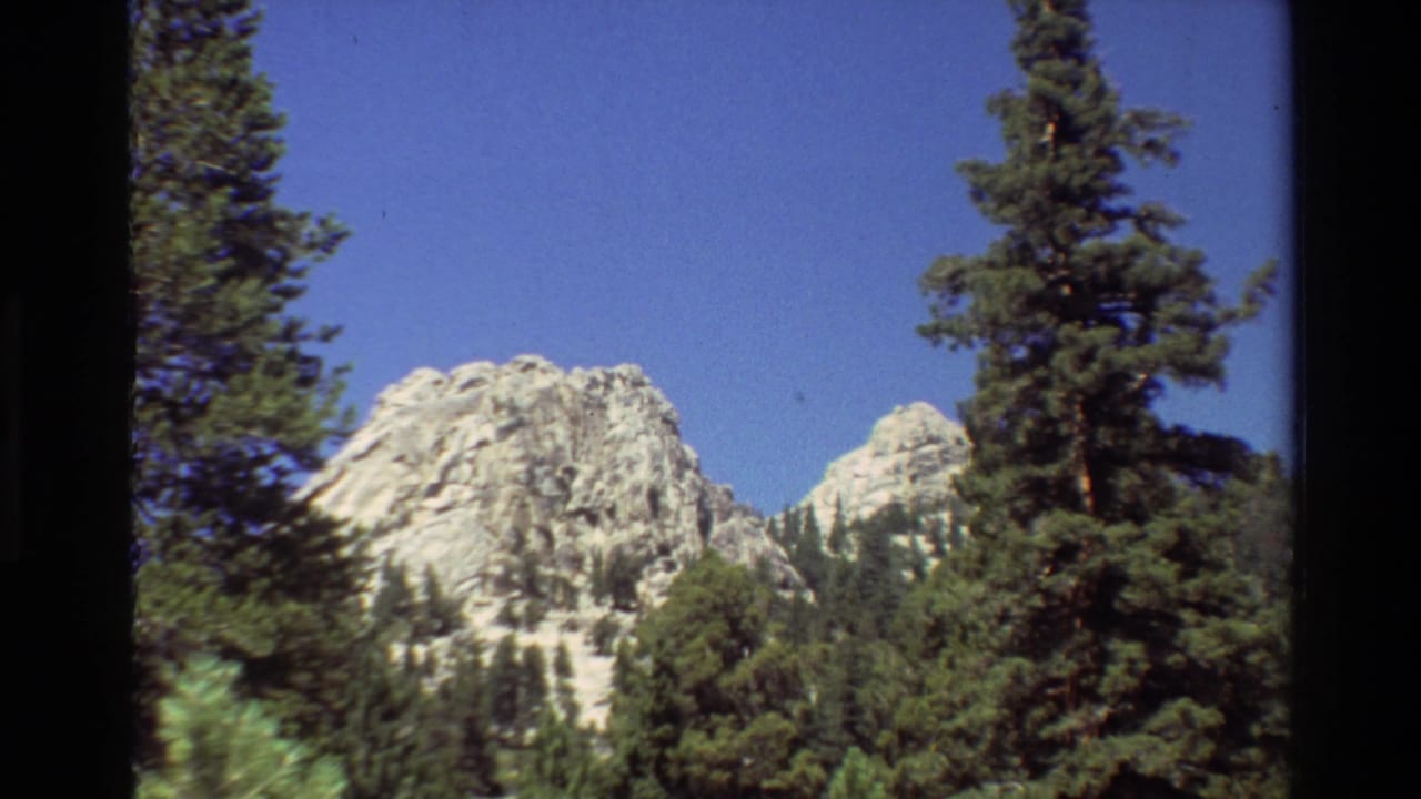 1981 8mm home-movie frame of a pale rocky mountain face seen through tall evergreens under clear sky.
