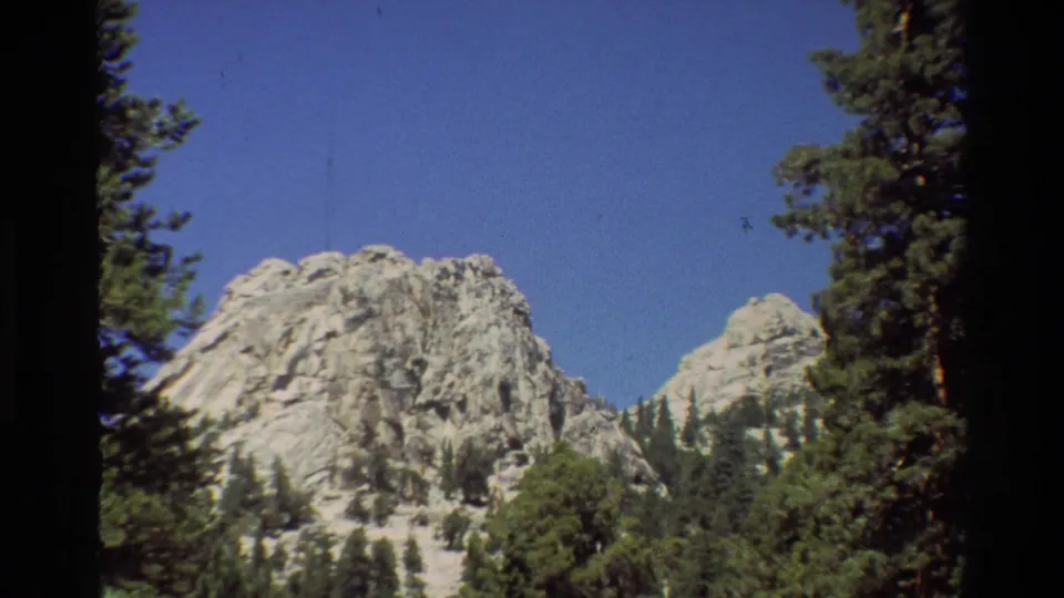 8mm frame of a pale mountain face partly lowered in view, with dense evergreens across the foreground.