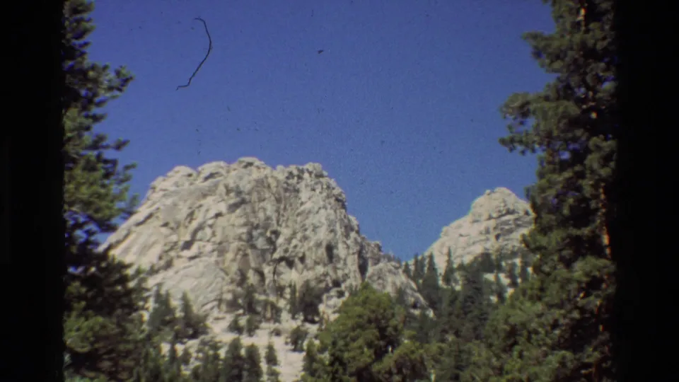 1981 8mm frame of a rocky mountain behind evergreens, with a small scratch visible against the blue sky.