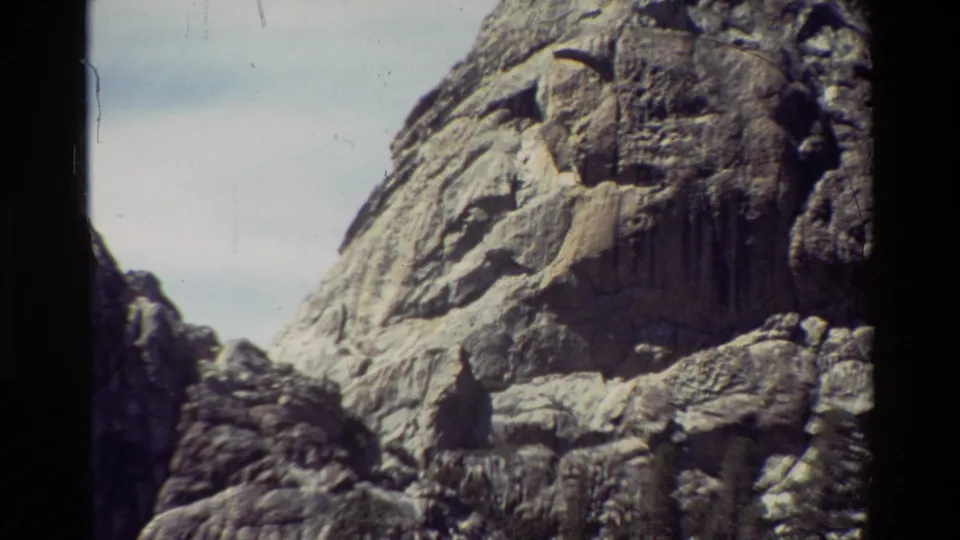 Later 8mm frame of the same rocky mountain face, with pale sky above and dark foreground boulders below.