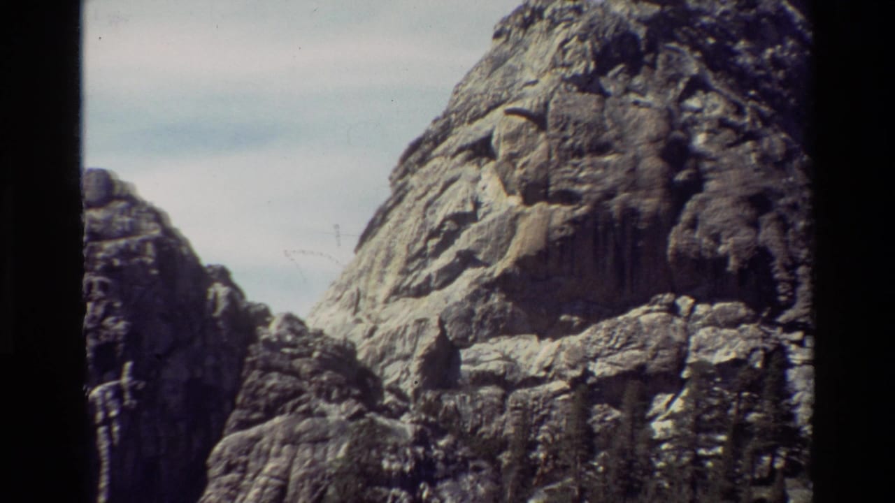 1981 8mm frame of a rugged rocky peak with smaller cliffs, pale sky, and visible film gate borders.